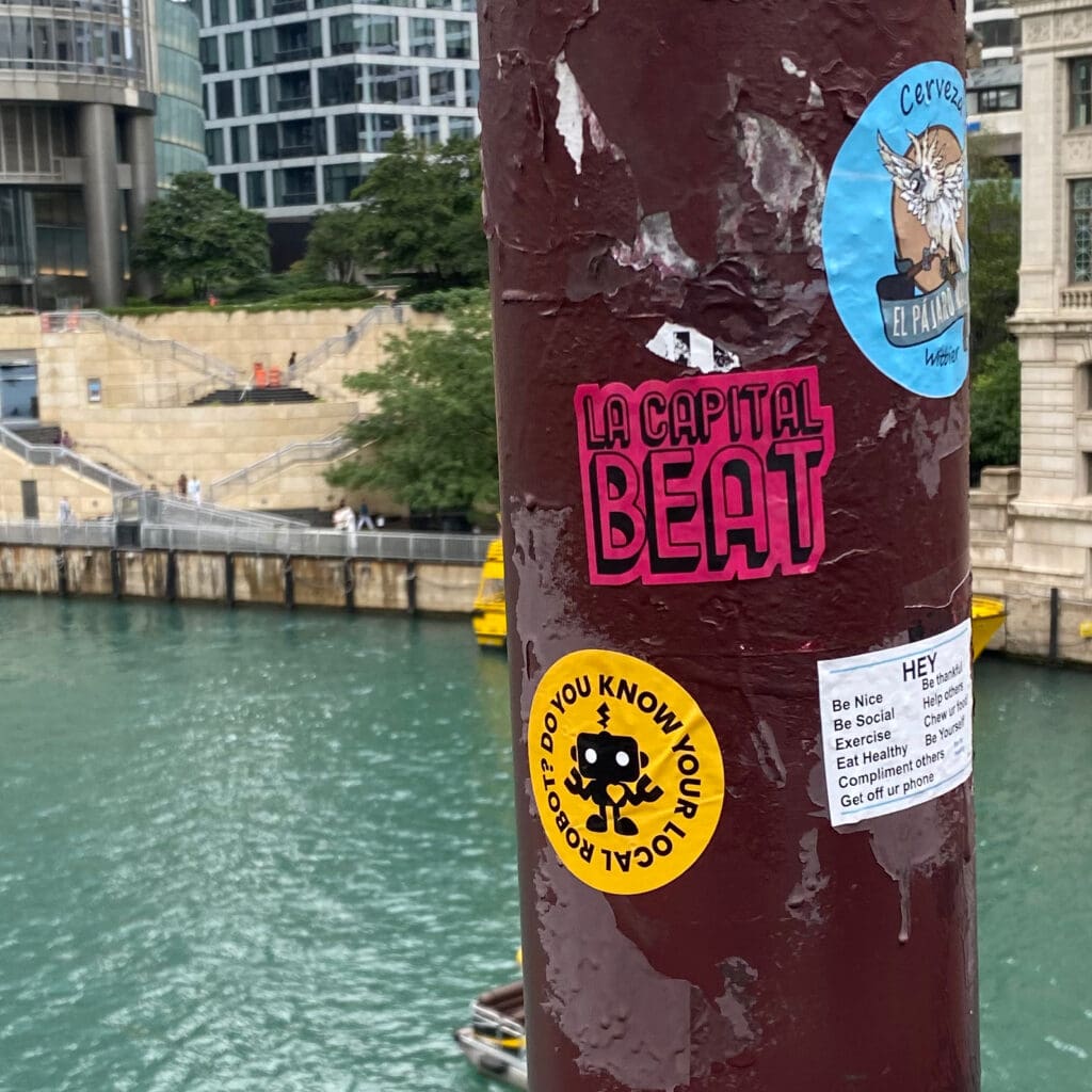 Stickers on a pole by a river: "Do You Know Your Local Robot?" sticker