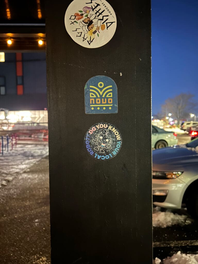Stickers on pole at night: "Do You Know Your Local Robot?" Robot graphic, snowy street background.
