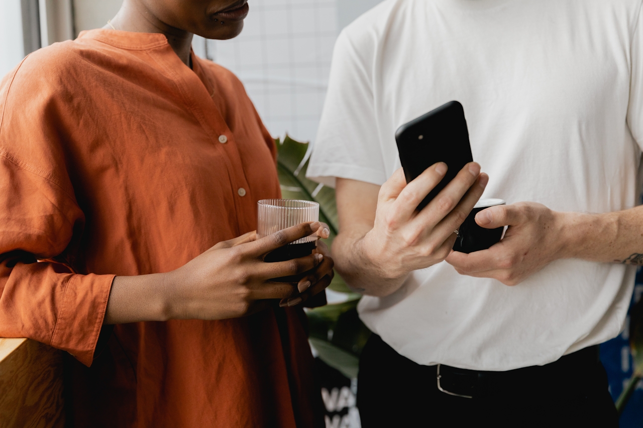 Two people engage in conversation, one holding a phone and the other a glass. Green plants are in the background.
