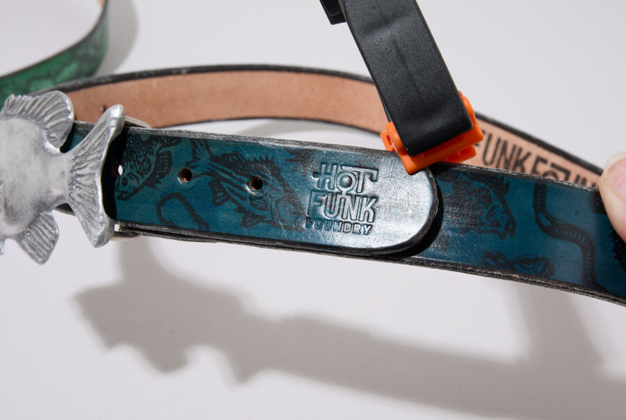 A hand holds a belt featuring a fish-shaped buckle and "Hot Funk Foundry" branding on a blue leather strap.